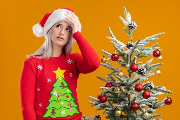 Jeune femme blonde en pull de Noël et bonnet de Noel jusqu'à confondu avec la main sur sa tête pour erreur