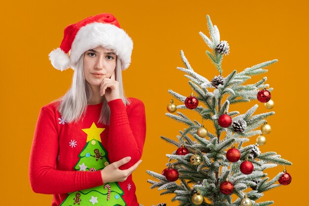 Jeune femme blonde en pull de Noël et bonnet de noel avec la main sur la pensée du menton