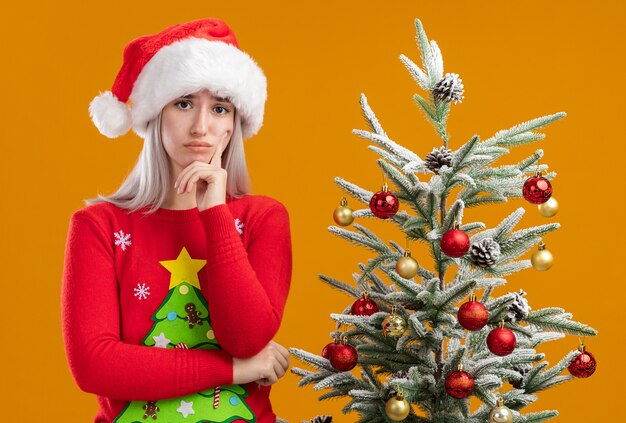 Jeune femme blonde en pull de Noël et bonnet de Noel regardant la caméra perplexe debout à côté d'un arbre de Noël sur fond orange