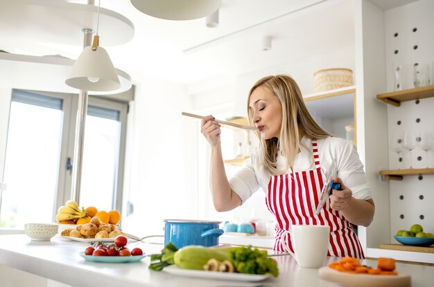 Jeune femme blonde de race blanche avec un tablier à rayures rouges dégustation de soupe à l'aide d'une cuillère en bois dans une cuisine