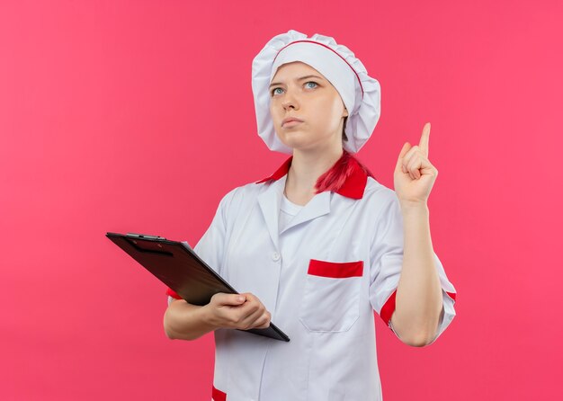 Jeune femme blonde réfléchie chef en uniforme de chef tient le presse-papiers et pointe vers le haut isolé sur mur rose