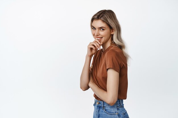 Jeune femme blonde séduisante riant et souriant, coning la bouche avec la main, regardant coquette, debout sur blanc