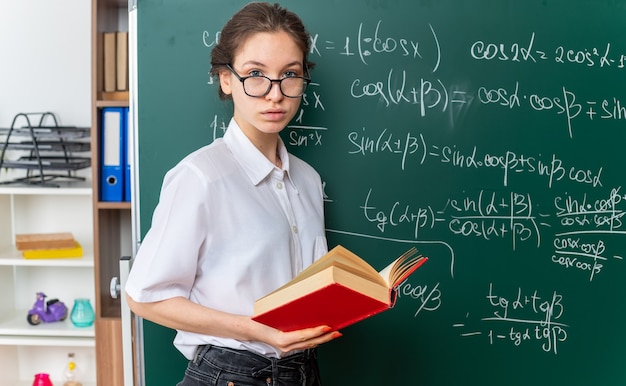 Jeune femme blonde sérieuse professeur de mathématiques portant des lunettes debout dans la vue de profil devant le tableau tenant un livre en classe