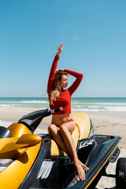 Jeune femme blonde sexy avec un corps parfait en tenue rouge assis sur un scooter des mers sur la plage sous un soleil. Week-end d'été ou vacances. Sport extrême.