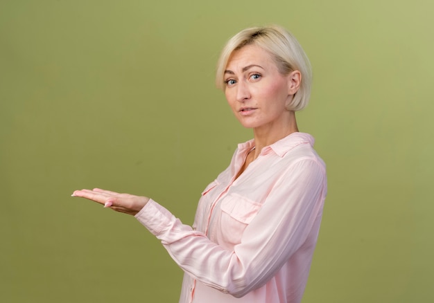 Jeune femme blonde slave points avec la main à côté isolé sur mur vert olive avec espace copie