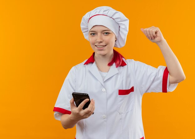 Jeune femme blonde souriante chef en uniforme de chef tient le téléphone et lève le poing isolé sur mur orange