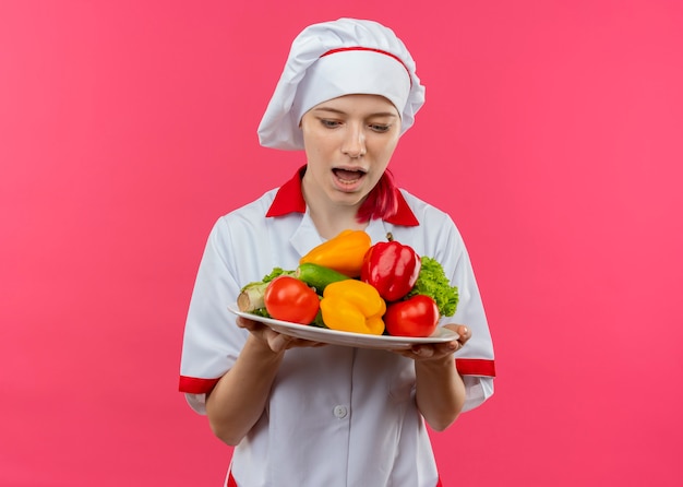 Jeune femme blonde surprise chef en uniforme de chef détient des légumes sur une plaque isolée sur un mur rose