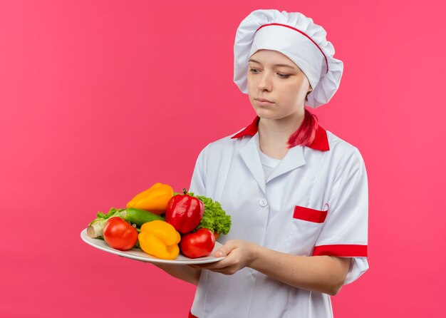 Jeune femme blonde surprise chef en uniforme de chef tient et regarde les légumes sur la plaque isolée sur le mur rose