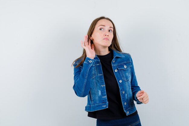 Jeune femme en blouse entendant une conversation privée et ayant l'air curieuse, vue de face.