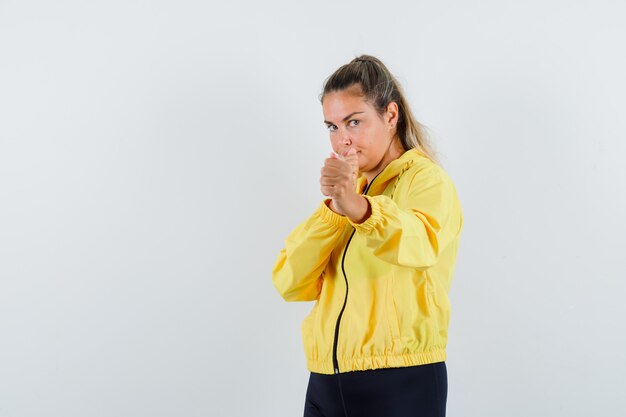 Jeune femme en blouson aviateur jaune et pantalon noir debout dans la pose de boxeur et à la sérieuse