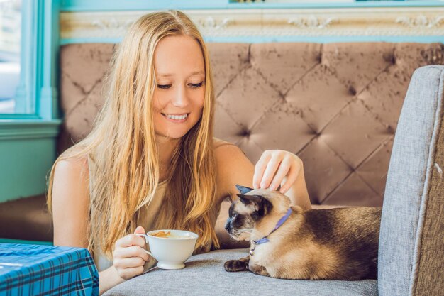La jeune femme boit du café et caresse le chat dans le contexte du canapé rayé par les chats