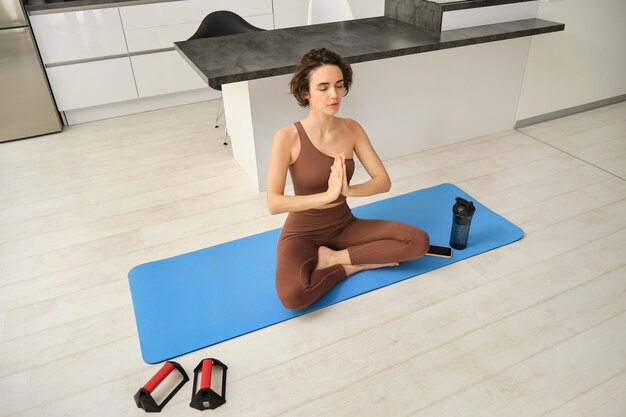 Une jeune femme en bonne santé et calme portant des vêtements de sport est assise à la maison dans le salon en train de faire du yoga.