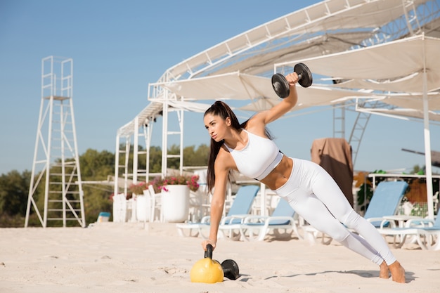 Photo gratuite jeune femme en bonne santé, formation du haut du corps avec des poids à la plage.