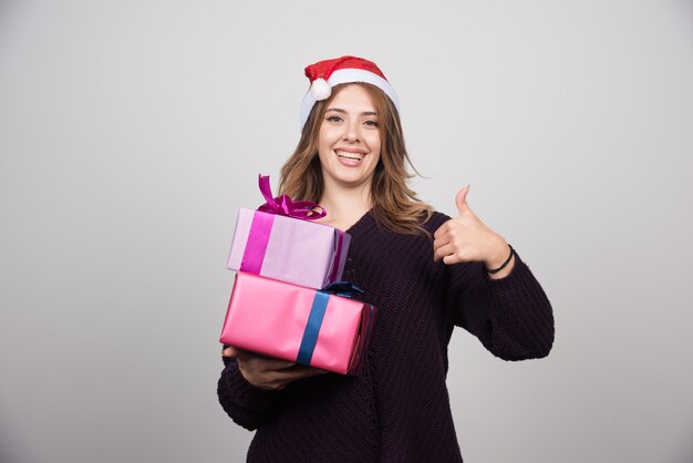 Jeune femme en bonnet de Noel avec des coffrets cadeaux montrant un pouce vers le haut.