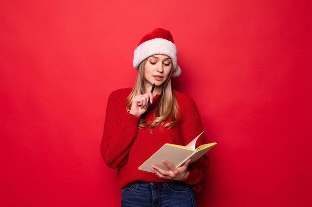 Une jeune femme avec un bonnet de Noel tient un bloc-notes dans ses mains et mord un stylo en vérifiant la liste des cadeaux ou des enfants ayant un bon comportement isolés sur un mur rouge.,