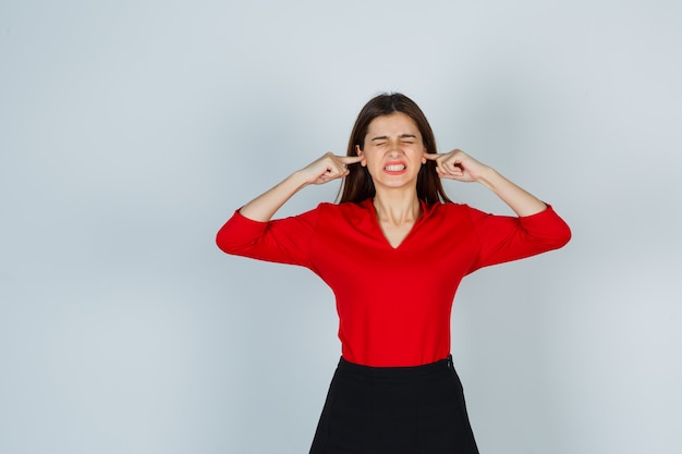 Jeune femme bouchant les oreilles avec les doigts en chemisier rouge, jupe et à l'ennui