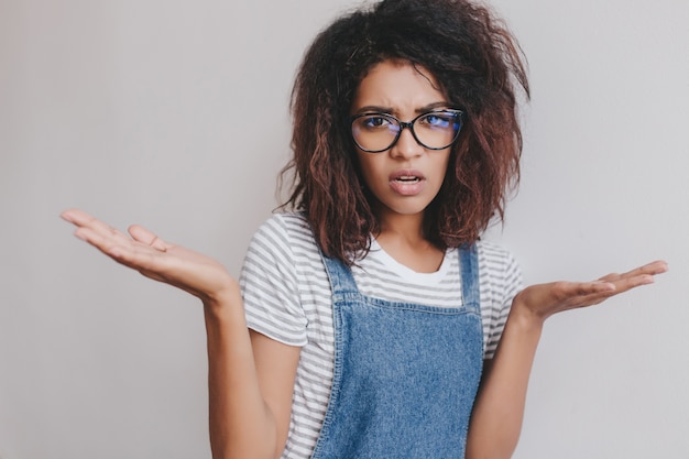 Jeune femme bouclée à la peau brun clair debout avec les paumes vers le haut et l'expression du visage déplacé