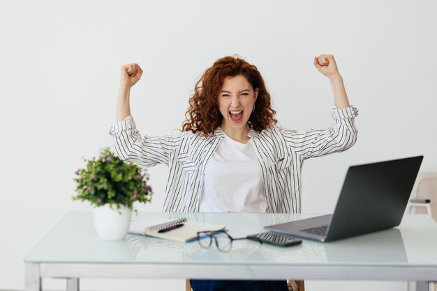 Jeune femme bouclée rousse travaillant avec son ordinateur portable levant le poing après un concept de vainqueur de la victoire