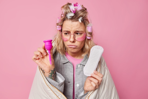 Une Jeune Femme Bouleversée Choisit Entre Une Coupe Menstruelle Et Une Serviette Hygiénique A De La Mauvaise Humeur Pendant La Menstruation Porte Des Lunettes De Pyjama Enveloppées Dans Une Couverture Applique Des Tampons D'hydrogel Sous Les Yeux Pose à L'intérieur