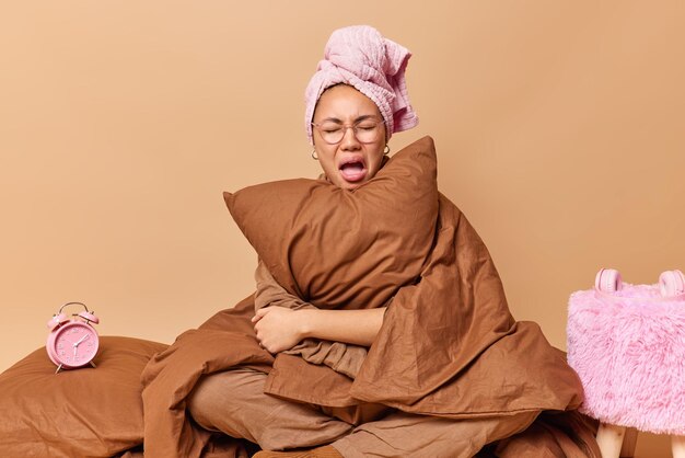 Une jeune femme bouleversée pleure de désespoir enveloppée dans une couverture embrasse l'oreiller porte une serviette sur la tête et le pyjama est assis les jambes croisées dans le lit se sent malheureuse d'entendre la sonnerie du réveil isolé sur un mur beige