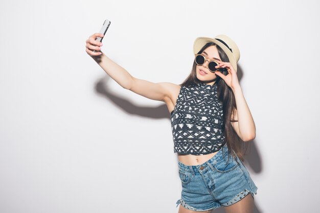 Jeune femme brune attrayante joyeuse sourit sur le mur blanc prenant selfie avec téléphone, portant une tenue d'été décontractée et un chapeau