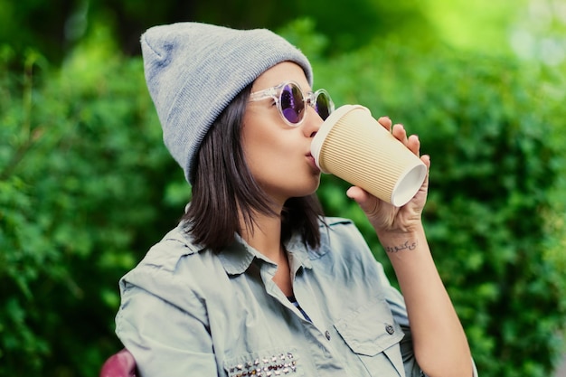 Une jeune femme brune au chapeau boit du café dans une tasse en papier dans un parc verdoyant d'été.