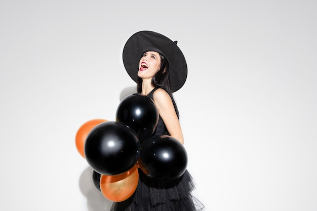 Jeune femme brune au chapeau noir et costume sur fond blanc. Modèle féminin caucasien attrayant. Halloween, vendredi noir, cyber lundi, ventes, concept d'automne. Copyspace. Contient des ballons, effrayant.