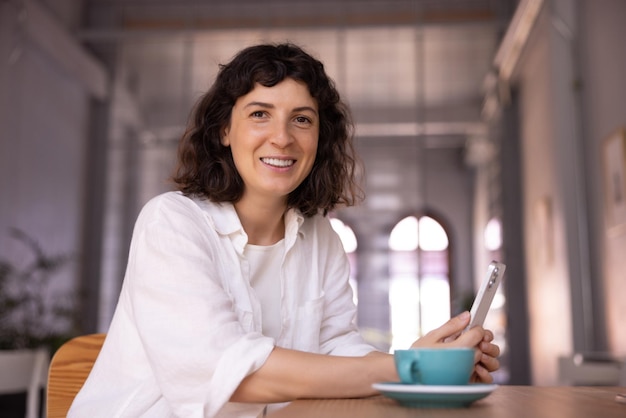 Jeune femme brune caucasienne moderne en chemise blanche regarde la caméra et tient un smartphone assis à table avec du café