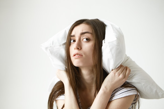 Photo gratuite jeune femme brune avec une coiffure en désordre couvrant les oreilles à l'aide d'un oreiller blanc à la recherche de côté avec frustration
