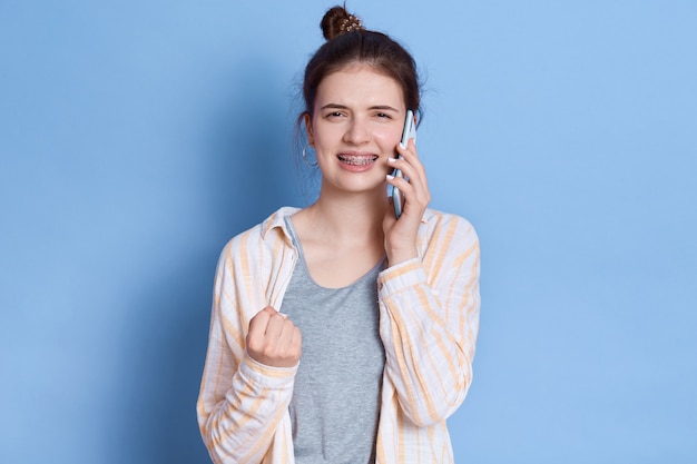 Jeune femme brune a une conversation avec un ami et un poing serré, vêtue d'une tenue décontractée, debout contre le mur bleu.