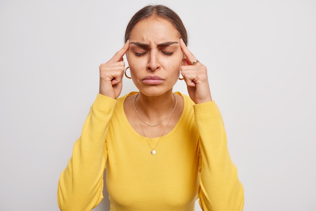 Une jeune femme brune frustrée garde l'index sur les tempes souffre de terribles maux de tête ou de migraine garde les yeux fermés pour révéler la douleur porte un pull jaune décontracté isolé sur fond blanc