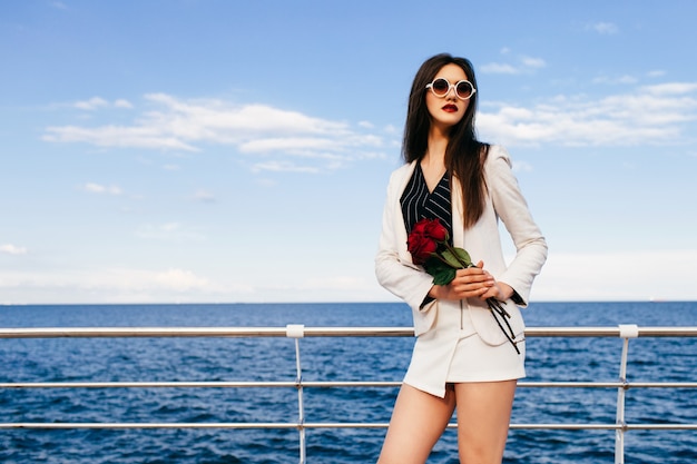 Jeune femme brune hipster posant au remblai avec petit bouquet rouge de roses