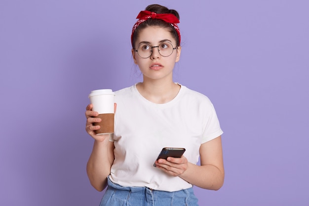 Jeune femme brune isolée sur espace lilas tenant du café à emporter et mobile tout en pensant à quelque chose