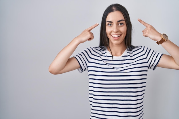 Jeune femme brune portant un t-shirt rayé souriant pointant vers la tête avec le doigt des deux mains, bonne idée ou pensée, bonne mémoire