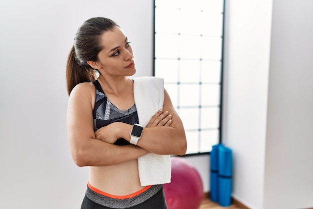 Jeune femme brune portant des vêtements de sport et une serviette à la salle de gym regardant sur le côté avec les bras croisés convaincu et confiant