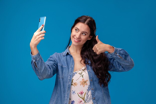 Jeune femme brune pose avec un smartphone debout sur fond bleu.