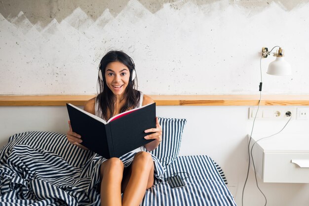 Jeune femme brune séduisante assise sur le lit en pyjama, souriant, chambre, émotion heureuse, se réveiller le matin, écouter de la musique sur les écouteurs, livre de lecture, tenant l'album, expression du visage riant