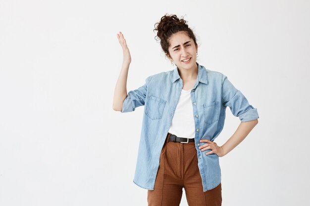 Une jeune femme brune serre les dents dans l'irritation vêtue d'une chemise en jean sur un haut blanc, étant ennuyée et insatisfaite du travail de ses collègues, faisant des gestes activement, gardant la main sur la hanche.