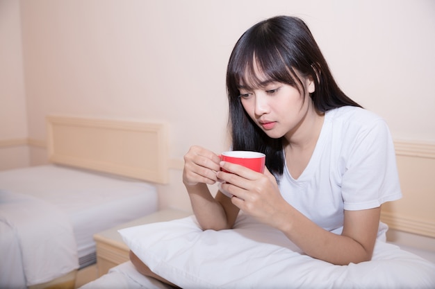 Jeune femme buvant du café à la maison dans son lit et vérifiant son ordinateur portable