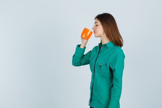 Jeune femme buvant du thé dans une tasse orange en chemise et à la vue de face, focalisée.
