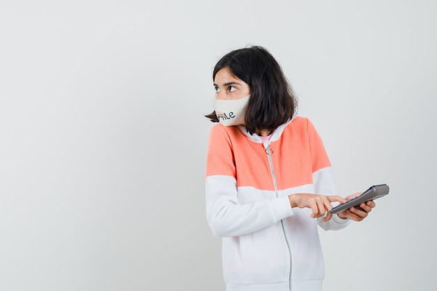 Jeune femme calculant avec calculatrice en sweat à capuche, masque facial et à la recherche concentrée.