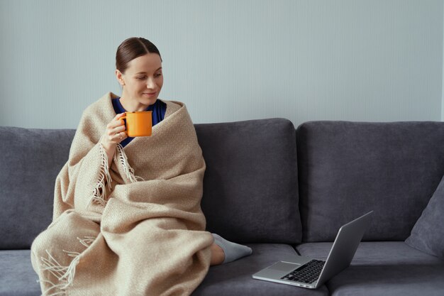 Jeune femme câlins dans une couverture chaude avec un ordinateur portable et une tasse de thé