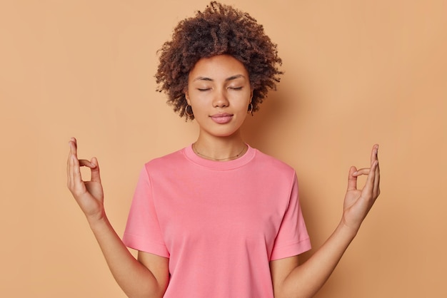 Une jeune femme calme et concentrée garde les yeux fermés les doigts ensemble médite les pratiques d'intérieur le yoga porte un t-shirt rose décontracté isolé sur fond marron essaie de trouver la paix se tient dans une pose zen