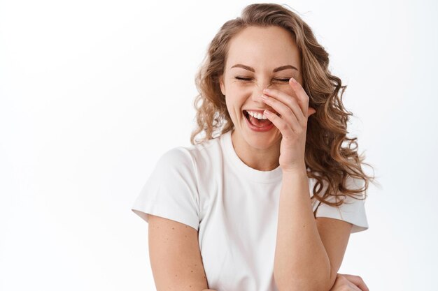 Jeune femme candide aux cheveux blonds, riant à haute voix, couvrant la bouche tout en souriant et en riant de quelque chose de drôle, entendant une blague hilarante, debout sur fond blanc