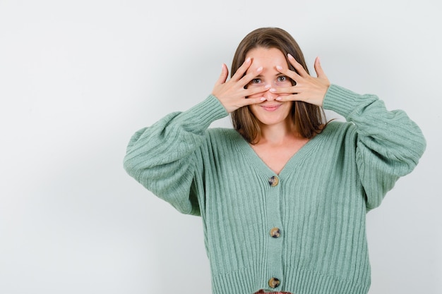 Photo gratuite jeune femme en cardigan en laine à la recherche à travers les doigts et à la jolie vue de face.