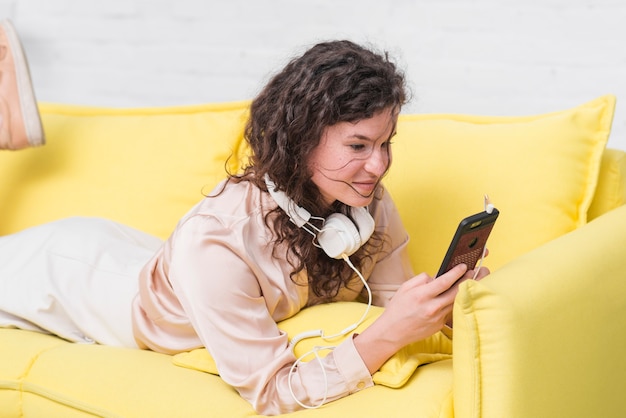 Jeune femme avec un casque autour du cou, couché sur un canapé jaune à l&#39;aide de smartphone