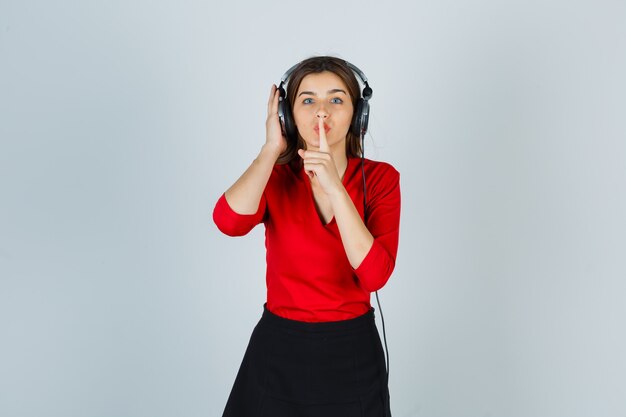 Jeune femme avec un casque d'écoute de la musique tout en montrant le geste de silence en chemisier rouge