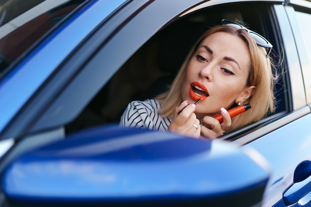 Jeune femme caucasienne, appliquer le rouge à lèvres en regardant la réflexion dans le miroir de la voiture.