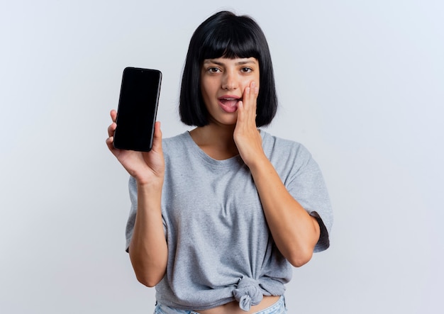 Une jeune femme caucasienne brune excitée met la main sur le visage et tient le téléphone