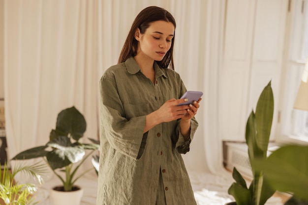 Une jeune femme caucasienne calme regarde l'écran du téléphone portable dans ses mains à l'intérieur Une brune en longue chemise verte lit des SMS sur le réseau social Concept d'appareil technologique